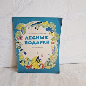 Op de afbeelding: Een vintage kleurboek uit 1978 met de titel "ЛЕСНЫЕ ПОДАРКИ" en een blauwe rand. De omslag heeft een crèmekleurig middengedeelte versierd met illustraties van bosplanten, bessen, bloemen en een gele mand gevuld met paddenstoelen.