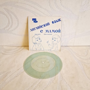 Könnte beinhalten: Ein Vintage-Kinderbuch mit dem Titel "Englisch mit Mama" auf Russisch, mit Illustrationen von zwei Kindern. Das Buch ist weiß mit blauem Text und Illustrationen. Eine durchscheinende grüne Schallplatte befindet sich im Vordergrund.