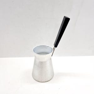 May include: A small, silver-colored coffee pot with a black handle. The pot has a conical shape, wider at the base and narrowing towards the top. The handle is rectangular and attached to the side of the pot.