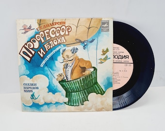 May include: A vintage vinyl record with a black label and a colorful illustration of a man in a green basket, floating in the sky. The illustration is titled "Professor and the Flea" and includes the text "Tales of the Peoples of the World".