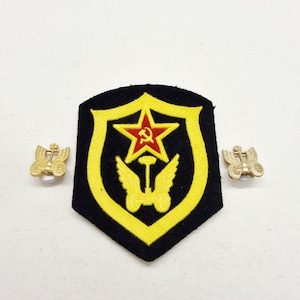 May include: A black and yellow patch with a red star and hammer and sickle emblem, flanked by two gold-colored pins. The patch features a shield design with a yellow border.