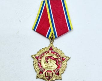 May include: A commemorative medal featuring a gold-toned five-pointed star with red enamel accents. The center depicts a soldier and tank with the text "КРАСНОЙ АРМИИ" and "100 ЛЕТ". It is suspended from a ribbon with red, yellow, blue, and white stripes.