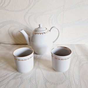May include: A white teapot with a gold floral design and two matching white teacups with gold floral designs.