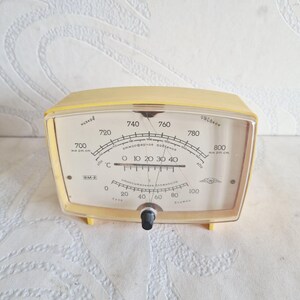 May include: A vintage yellow barometer with a clear plastic face. The gauge displays atmospheric pressure in mm Hg and temperature in Celsius. The face has black markings and a black knob at the bottom.