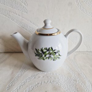 Op de afbeelding: Een witte porseleinen theepot met een delicate bloemenillustratie van groene en witte sneeuwklokjes op de voorzijde. De theepot heeft een bijpassend deksel met een goudkleurige rand en een gebogen handvat met een gladde, glanzende afwerking.