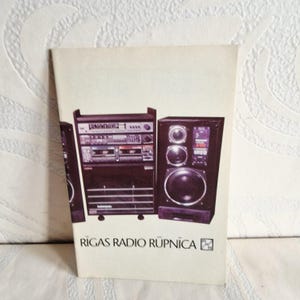 Könnte beinhalten: Eine Vintage-Werbung mit einer Stereoanlage mit schwarzem Schrank und Lautsprechern. Der Text "RIGAS RADIO RUPNICA" steht unten. Das Bild befindet sich auf weißem Hintergrund.