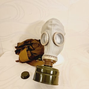Op de afbeelding: Een grijs gasmasker met twee ronde oogstukken en een groen filter. Het masker is bevestigd aan een bruin band en een bruine canvas tas.
