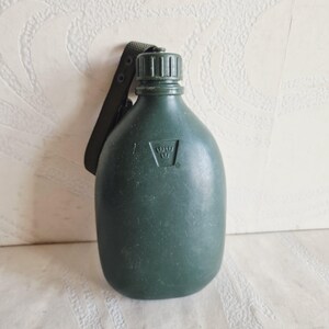 Pode incluir: Uma cantil verde escuro com uma tampa de rosca a condizer e uma correia preta. O cantil tem uma forma arredondada e um pequeno emblema em relevo. A correia é presa à tampa e à lateral do cantil.
