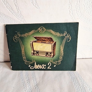 Puede incluir: Folleto vintage con fondo verde oscuro y elementos decorativos dorados. El folleto presenta una ilustración de una radio retro con el texto "Люкс 2". La radio es marrón y beige.