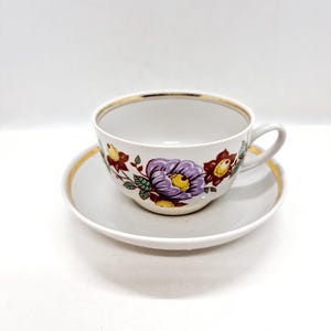 May include: A white porcelain teacup and saucer set with a floral pattern in shades of purple, red, yellow, and green. The cup and saucer have a gold trim.