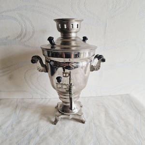 Puede incluir: Un samovar de metal plateado con un cuerpo central en forma de urna, una tapa extraíble con una chimenea ventilada y dos asas laterales. El recipiente incluye un grifo vertical con un pomo negro y detalles negros adicionales en la tapa y asas.