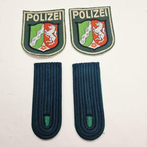Può includere: Un set di quattro elementi di uniforme della polizia vintage. Due toppe verdi con la scritta "POLIZEI" e uno stemma con un cavallo bianco. Due spalline blu scuro con dettagli verdi.
