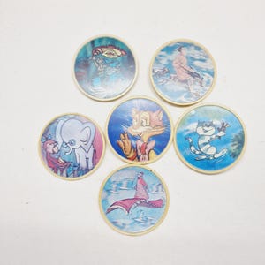 May include: Six round, colorful, plastic tokens with cartoon images. The tokens feature a cat, an elephant, a woman on a flying carpet, a cat riding a dog, a man riding a dog, and a man riding a tiger.
