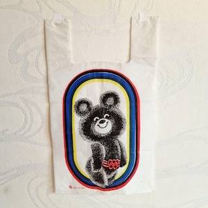 Op de afbeelding: Witte plastic boodschappentas met een zwarte berenillustratie in een kleurrijk ovaal frame. De beer heeft een lachend gezicht en draagt een rode riem met Olympische ringen. De tas heeft een handvat en is waarschijnlijk vintage.