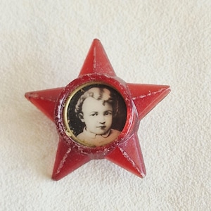 Könnte beinhalten: Eine vintage, sternförmige rote Brosche mit einem kleinen kreisförmigen Porträt eines Kindes. Das Schwarz-Weiß-Porträt ist in einem goldfarbenen Rahmen eingefasst. Der Stern hat ein leicht durchscheinendes Aussehen.