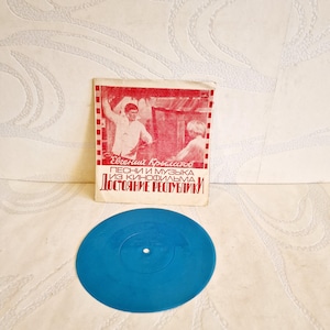 Könnte beinhalten: Eine blaue Schallplatte mit einem weißen Etikett. Die Schallplatte wird von einem kleinen, rot-weißen Papierumschlag mit dem Text "Евгений Кроматов Песни и музыка из кинофильма Достояние республики" begleitet.