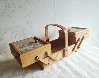 May include: A light-colored wooden sewing box featuring a cantilevered design with multiple tiered compartments, a central arched handle, and two lids with a decorative floral pattern inlay, connected by metal hinges and wooden support arms.