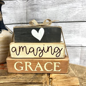 May include: Stacked wooden blocks with the words "amazing grace" and a white heart painted on the top block. The blocks are tied together with twine and a bow.