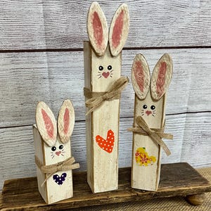May include: Three wooden bunny figures with cream-colored bodies, pink-lined ears, and painted faces. Each bunny has a jute twine bow and a heart design. The bunnies are displayed on a wooden stand, perfect for Easter decor.
