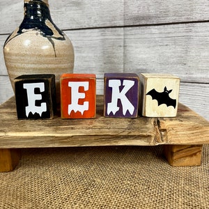 May include: Four wooden blocks spelling out "EEK" with a black bat on the last block. The blocks are painted in black, orange, purple, and white.