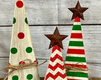 May include: Set of three wooden Christmas trees with rustic metal star toppers. The trees feature festive designs: polka dots, chevron, and stripes in red and green. Each tree is wrapped with twine.