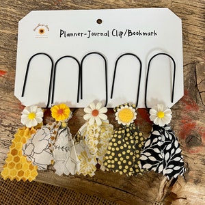 May include: A set of five black metal planner clips with fabric tassels and daisy accents. The fabric tassels feature yellow and white patterns, including honeycomb, floral, and polka dots. The card reads "Planner-Journal Clip/Bookmark."