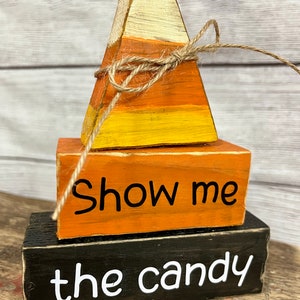 May include: A wooden tiered Halloween decoration with a candy corn shape on top. The bottom tier is black with white text that says "the candy". The middle tier is orange with white text that says "Show me".