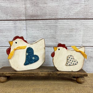May include: Two wooden chicken figurines with red combs, yellow beaks, and yellow ribbon bows. Each chicken has a blue heart with white polka dots. They sit on a rustic wooden stand, perfect for farmhouse decor.