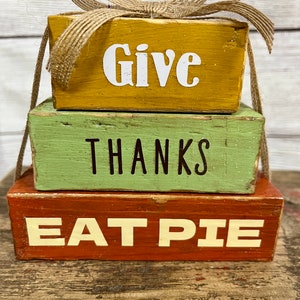 May include: A stack of three wooden blocks with the words "Give", "Thanks", and "Eat Pie" painted on them. The top block has a brown burlap bow.