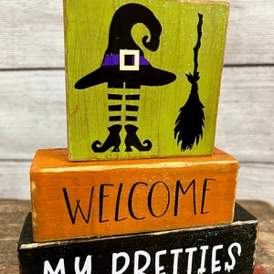 May include: A three-tiered wooden sign with a black witch silhouette, a purple hat, and a broom. The middle tier reads "WELCOME" in black lettering on an orange background. The bottom tier reads "MY PRETTIES" in white lettering on a black background.