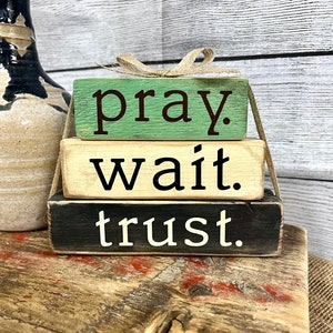 May include: Three wooden blocks stacked on top of each other. The top block is green and says "pray." The middle block is beige and says "wait." The bottom block is black and says "trust."