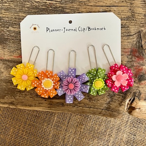 May include: Set of five colorful flower-shaped binder clips with white polka dots. The clips are attached to a white card with the text "Planner-Journal Clip/Bookmark".