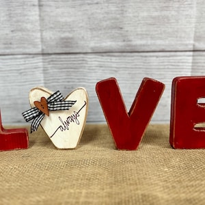 May include: Wooden LOVE sign with distressed red letters. The letter "O" is replaced with a wooden heart with a black and white checkered bow and the word "always" written on it. The sign is on a burlap surface.