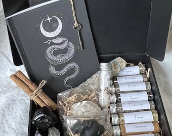May include: A black box filled with various spiritual items. Includes a notebook with a snake and crescent moon design, crystals, cinnamon sticks, small glass vials, and a key. The vials are labeled with different herbs.