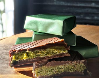 May include: Close-up of a layered dessert with a chocolate top and a green filling, possibly pistachio. Several wrapped green packages are stacked in the background. The dessert is cut to reveal the layers.