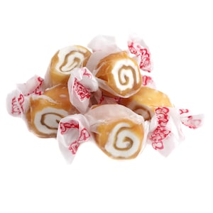 May include: A pile of caramel-colored candies with a white swirl center, individually wrapped in white paper with a red floral pattern. The candies have a spiral design and are a light brown color.