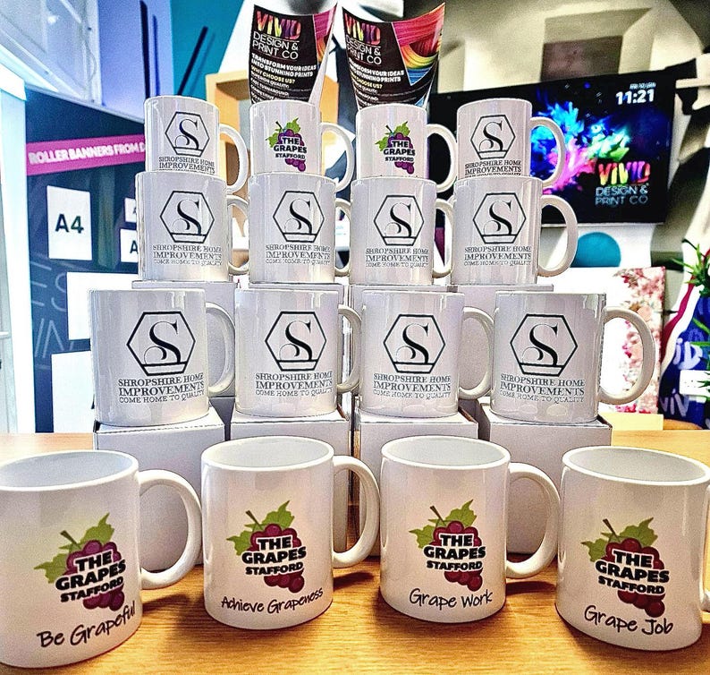 K&ouml;nnte beinhalten: Eine Sammlung wei&szlig;er Keramikbecher mit verschiedenen Designs. Einige Becher zeigen den Text "Shropshire Home Improvements" und einen stilisierten Buchstaben "S". Andere Becher zeigen den Text "The Grapes Stafford" mit einem Traubendesign und einem Slogan. Die Slogans lauten "Be Grapeful", "Achieve Grapeness", "Grape Work" und "Grape Job".
