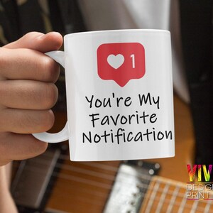 Può includere: Una tazza in ceramica bianca con un cuore rosso e il numero 1 in un fumetto. Il testo "You're my favorite notification" è stampato sulla tazza.