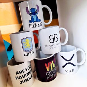 K&ouml;nnte beinhalten: Sechs wei&szlig;e Keramikbecher mit verschiedenen Designs. Die Becher sind &uuml;bereinander gestapelt. Die Designs beinhalten einen Comic-Charakter, ein Logo, ein Fu&szlig;ball-Wappen, ein Textdesign, ein Regenbogen-Design und ein Kryptow&auml;hrungs-Logo. Der Text auf den Bechern lautet "Tilly-Mae", "Baugh's Beauty & Boutique", "Aston Villa Giles", "Are you having a jiggle", "Viv Design Print" und "XRP".