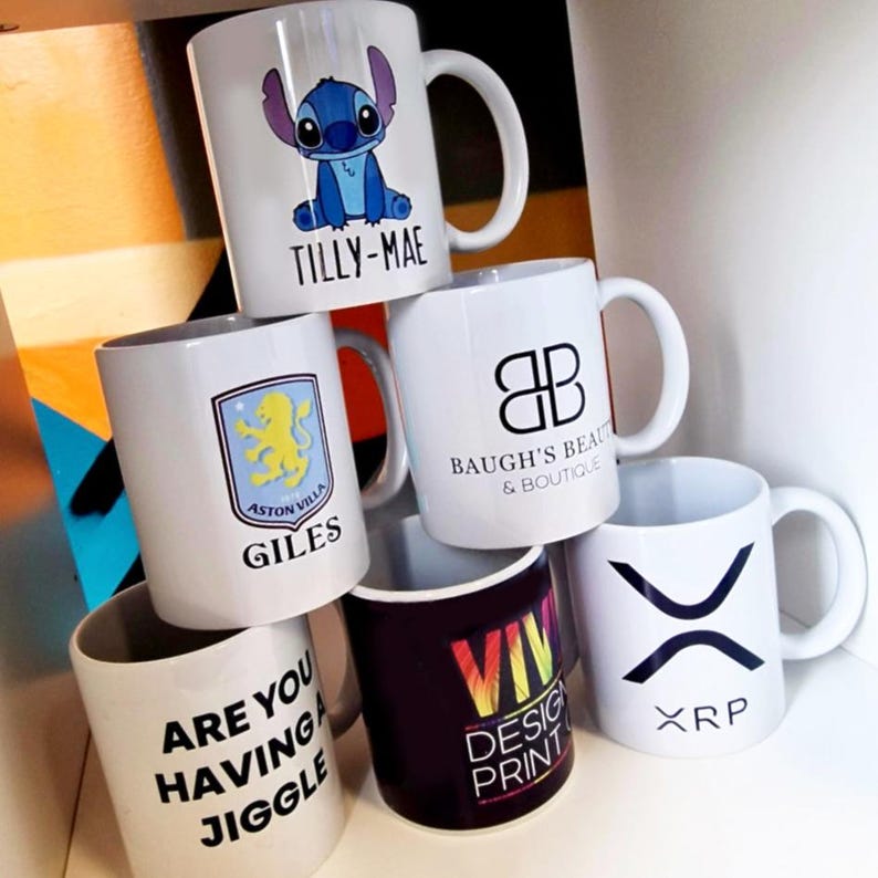 K&ouml;nnte beinhalten: Sechs wei&szlig;e Keramikbecher mit verschiedenen Designs. Die Becher sind &uuml;bereinander gestapelt. Die Designs beinhalten einen Comic-Charakter, ein Logo, ein Fu&szlig;ball-Wappen, ein Textdesign, ein Regenbogen-Design und ein Kryptow&auml;hrungs-Logo. Der Text auf den Bechern lautet "Tilly-Mae", "Baugh's Beauty & Boutique", "Aston Villa Giles", "Are you having a jiggle", "Viv Design Print" und "XRP".