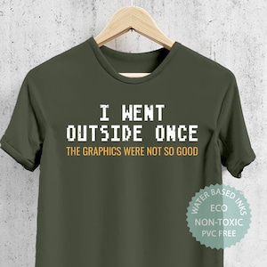 May include: A green t-shirt with the text "I WENT OUTSIDE ONCE THE GRAPHICS WERE NOT SO GOOD" in white pixelated font. The text is centered on the shirt. The shirt has a white tag on the bottom right corner that says "WATER BASED INKS ECO NON-TOXIC PVC FREE".