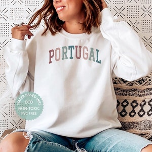 May include: White sweatshirt with the word "PORTUGAL" printed in colorful letters. The sweatshirt has a small, circular tag on the bottom left corner that says "Water Based Inks, Eco, Non-Toxic, PVC Free".