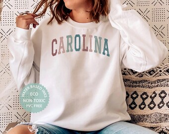 May include: A white sweatshirt with the word "CAROLINA" printed in rainbow colors. The sweatshirt has a small, round, green label with the text "WATER BASED INKS ECO NON-TOXIC PVC FREE".