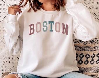Op de afbeelding: Een wit sweatshirt met het woord "BOSTON" bedrukt in kleurrijke letters. Het sweatshirt wordt gedragen door een persoon die op een bank zit met een wit deken.