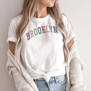 Op de afbeelding: Wit T-shirt met het woord "BROOKLYN" in een regenboogvormig ontwerp. Het shirt is in de taille geknoopt en wordt gedragen met een blauwe jeans en een cr&egrave;mekleurige cardigan.
