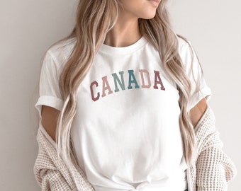 May include: A white t-shirt with the word "CANADA" printed in a rainbow color scheme. The shirt is tied at the waist and worn with a cream cardigan and blue jeans.