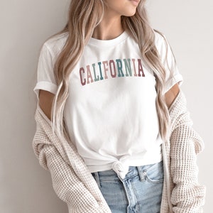 May include: A white t-shirt with the word "CALIFORNIA" printed in colourful letters. The letters are in shades of pink, blue, green, and brown.