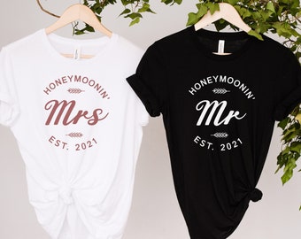 Puede incluir: Dos camisetas blancas con texto negro. Una camiseta dice "Honeymoonin' Mrs. Est. 2021" y la otra dice "Honeymoonin' Mr. Est. 2021".