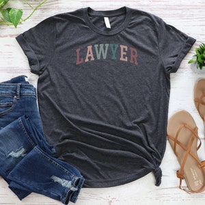 May include: A dark heather grey t-shirt with the word "LAWYER" printed in rainbow colors across the front.