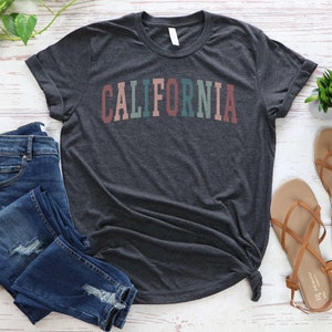 May include: A dark heather grey t-shirt with the word "CALIFORNIA" printed in rainbow colours across the front. The shirt is laid flat on a white wooden surface with a pair of blue jeans and brown sandals.